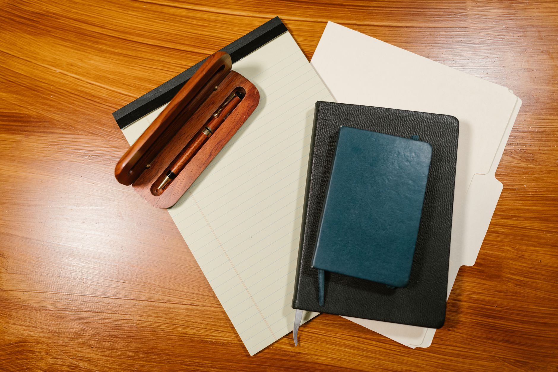 A pen case, a notepad, a notebook and a folder on a wooden desk, seen from above.