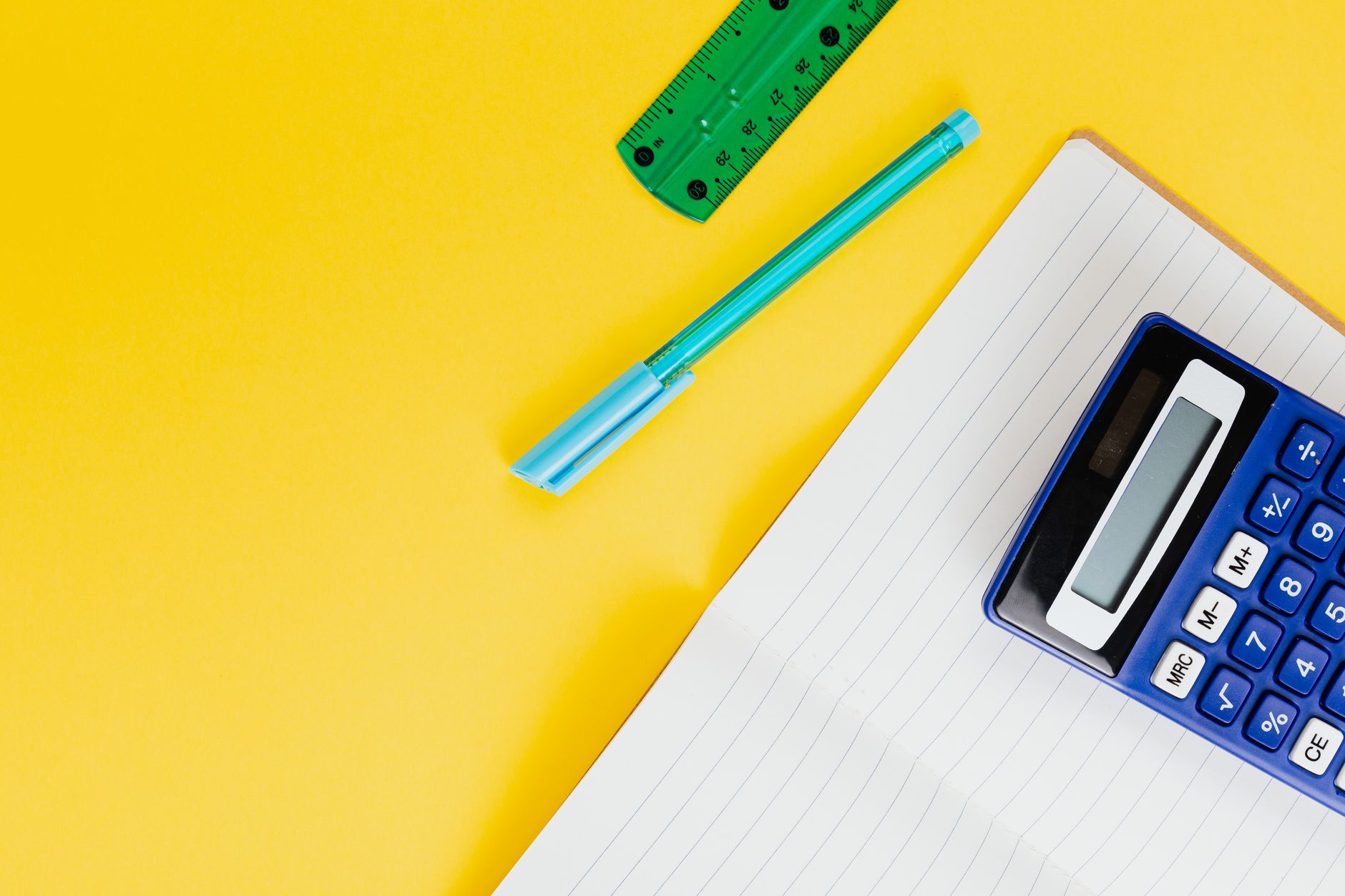 A blue calculator on a lined notebook beside a pen and a ruler on a yellow surface.