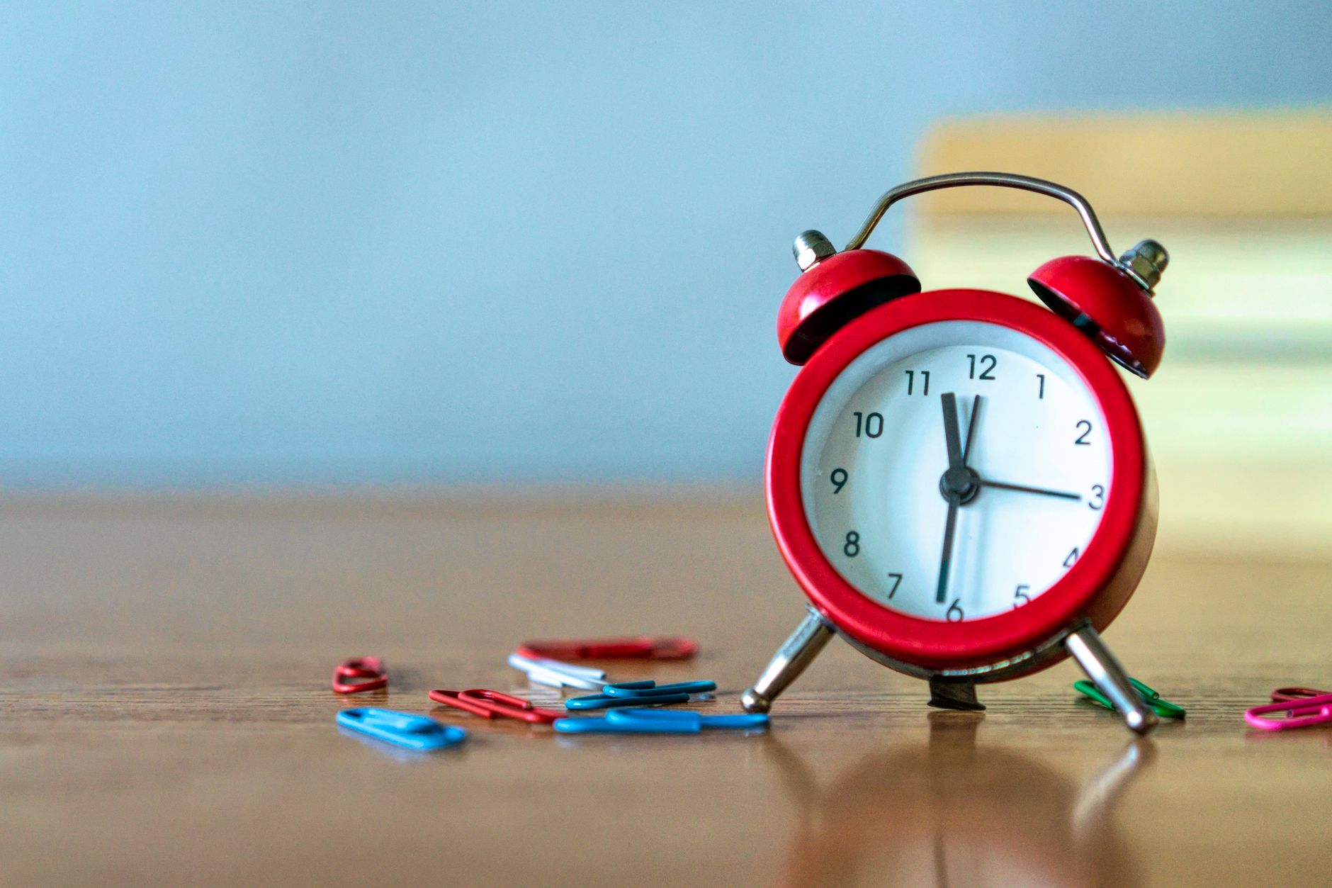 A red alarm clock and coloured paper clips on a wooden desk.