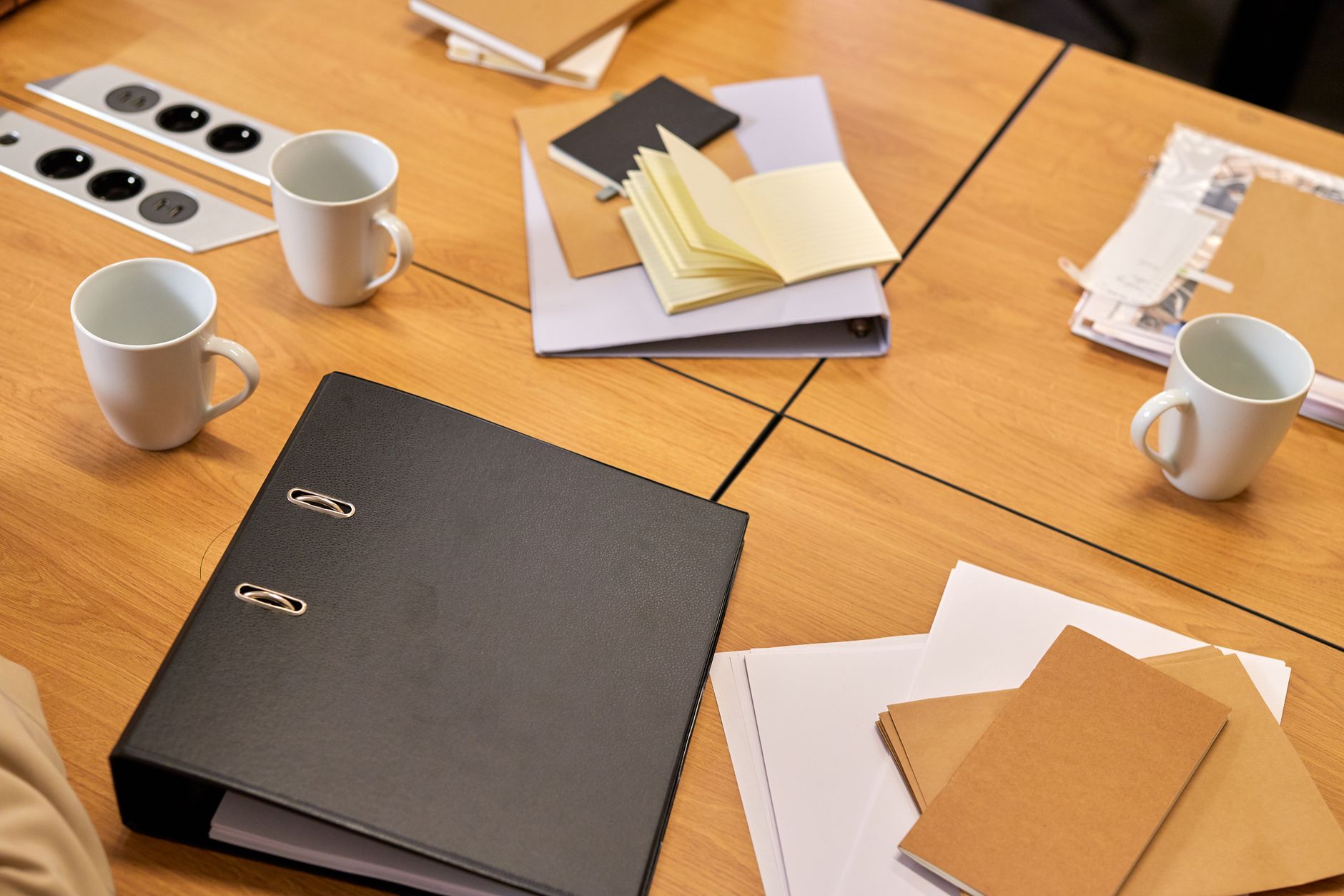 A black ring binder, loose papers and coffee mugs on a wooden office desk.