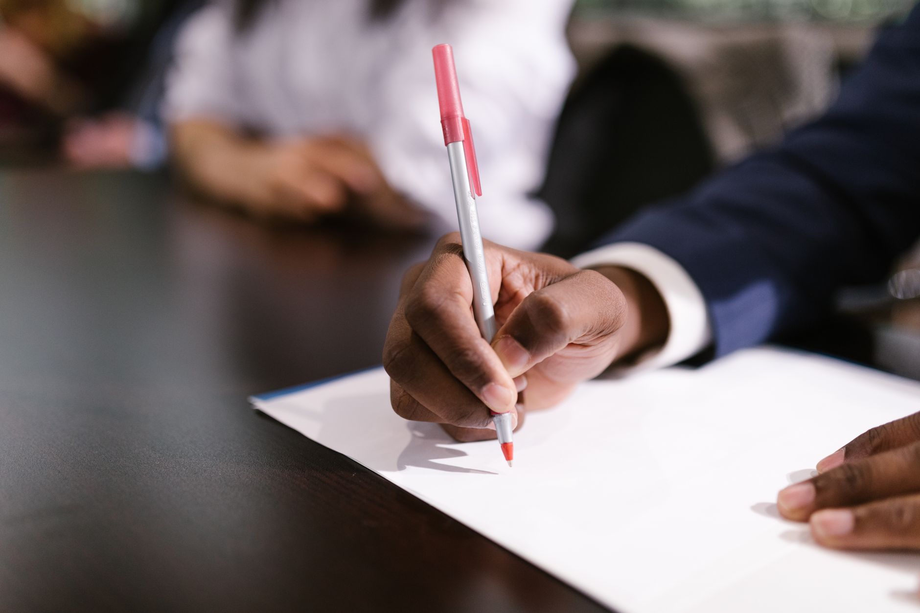 A hand writing on a sheet of white paper with a pen at a desk.