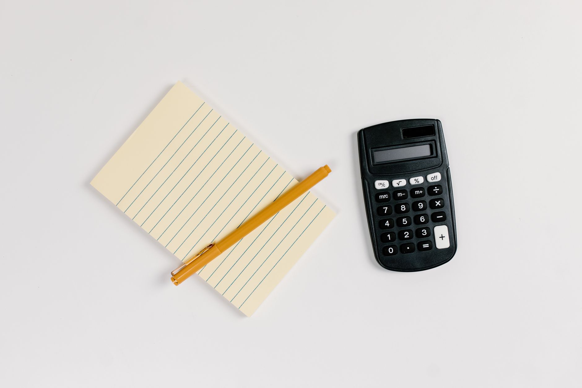 A calculator, a lined notepad and a pen on a white surface, seen from above.