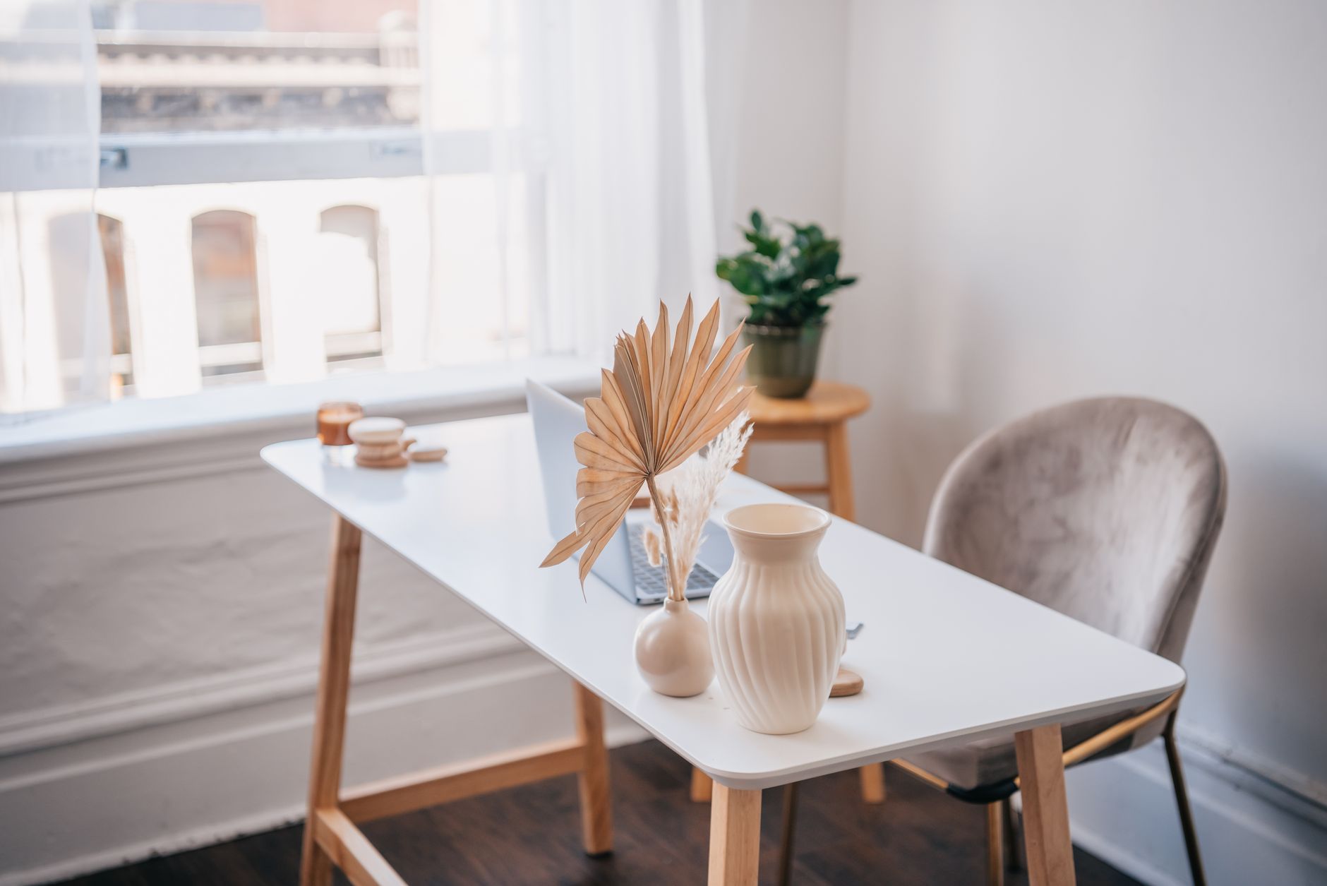 A white desk by a window with a laptop, a vase and a dried palm leaf.
