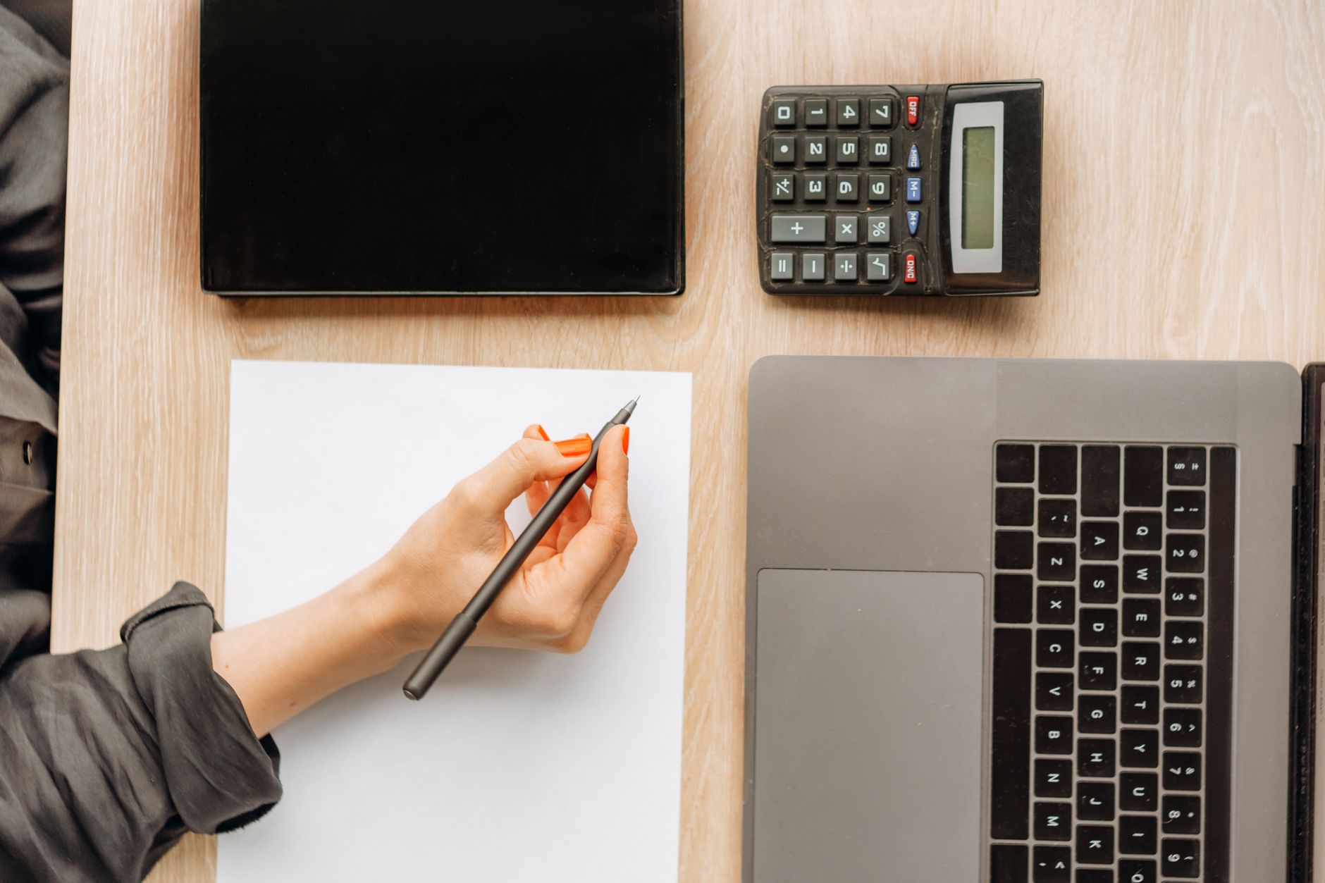 A hand writing on blank paper beside a laptop, a tablet and a calculator on an office desk, seen from above.