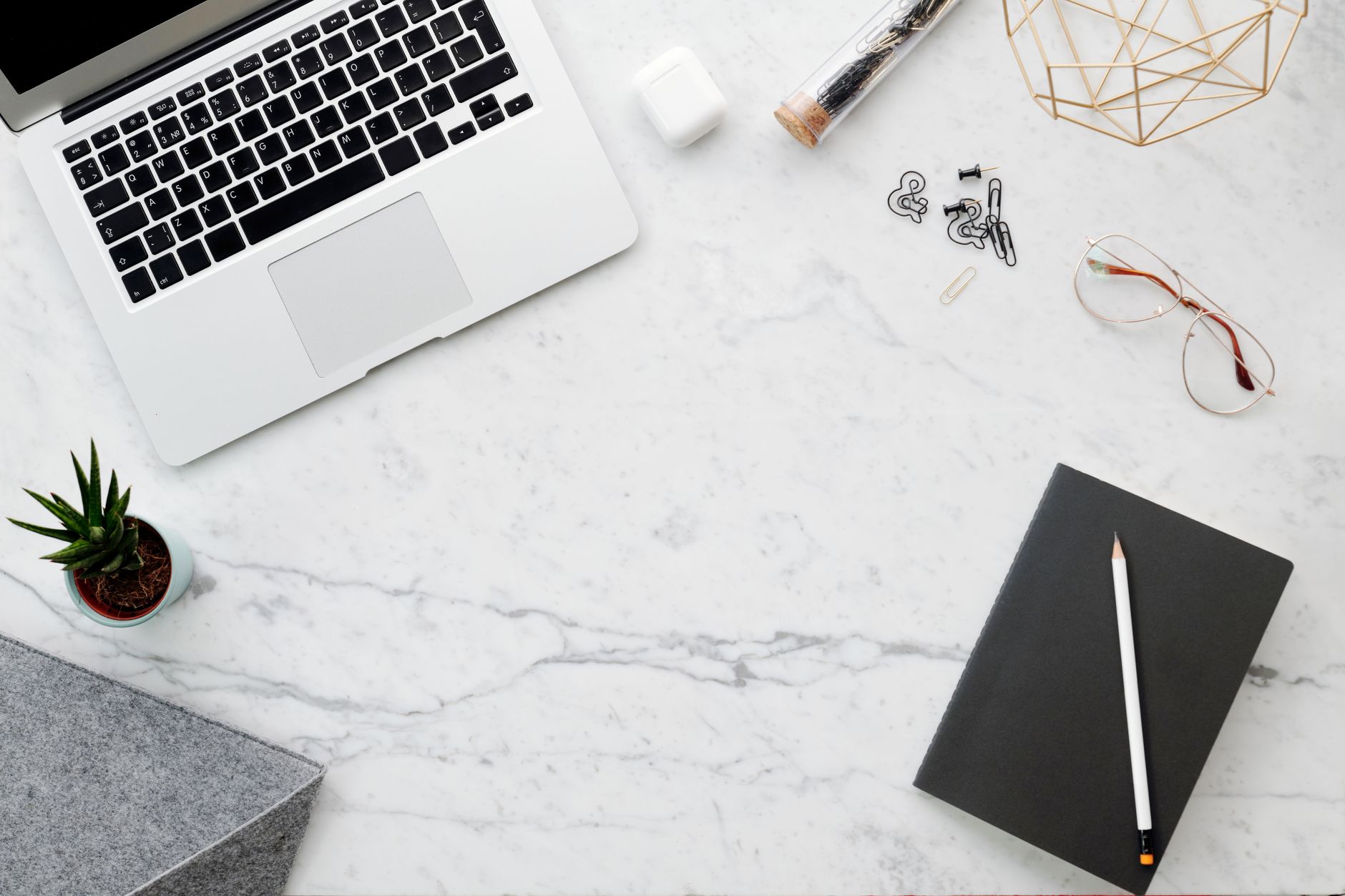A laptop, a notebook with a pencil, glasses and a small plant on a marble desk, seen from above.